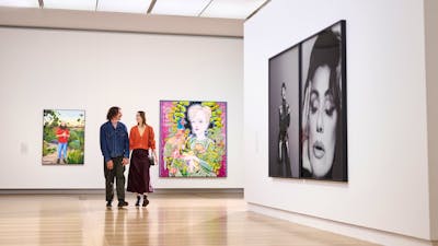 A couple is walking happily through a brightly lit gallery featuring several large portraits.