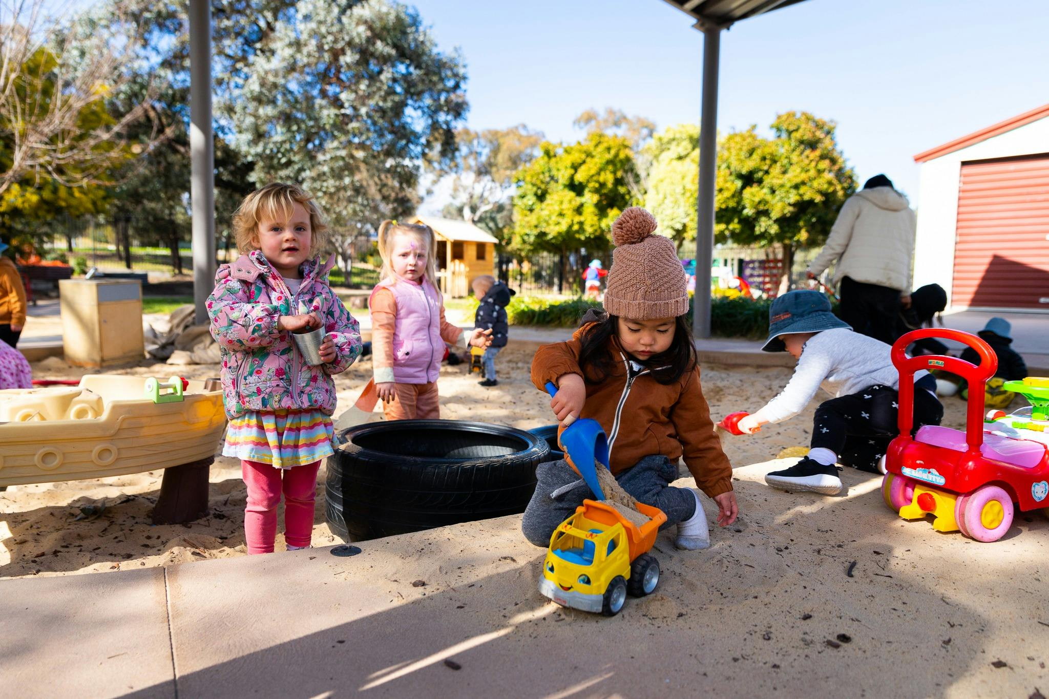 Young children play in a sandpit and look at the camera.
