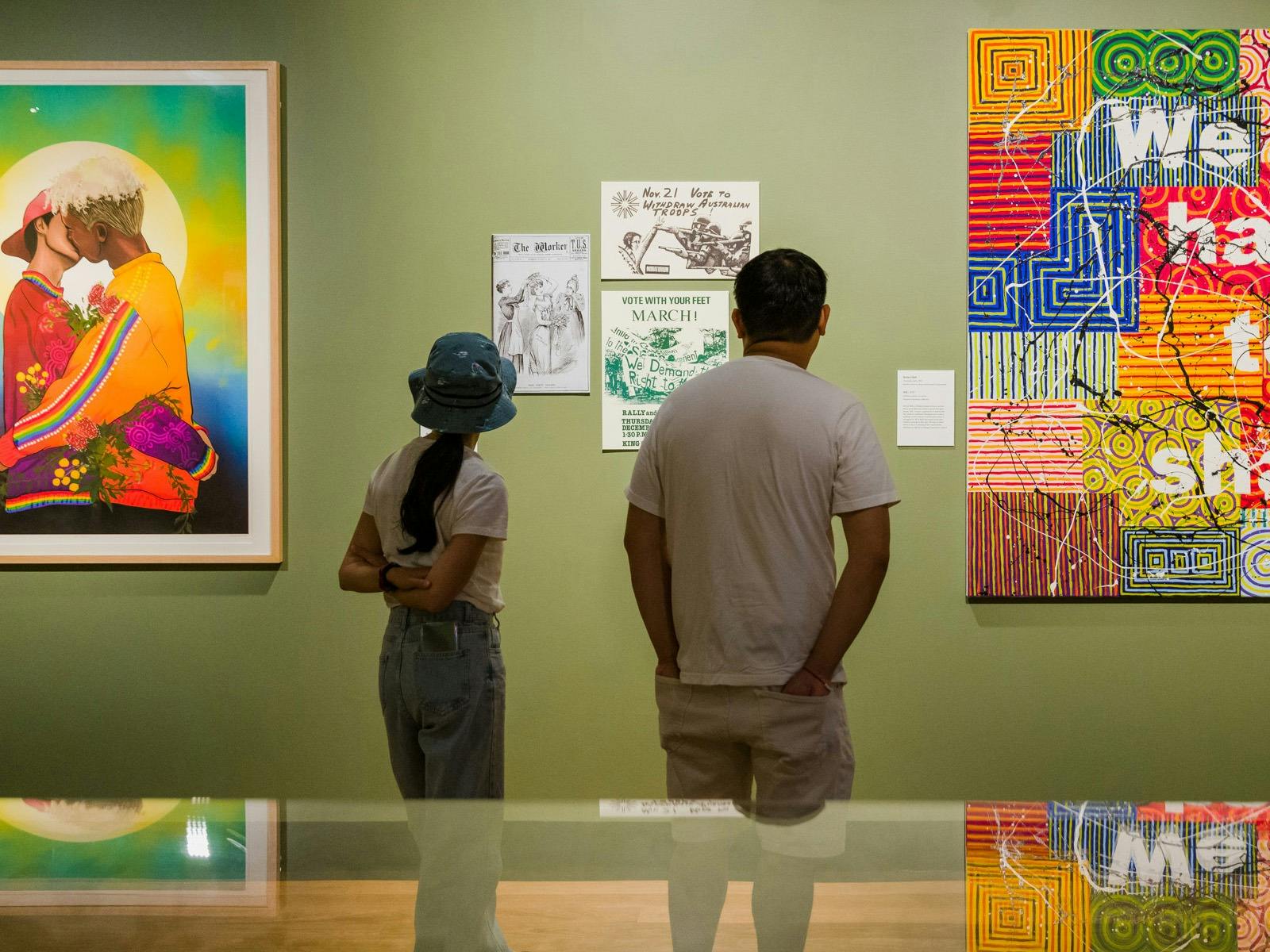 Two people stand in a green-walled gallery space with paintings on the walls. Two people stand in a green-walled gallery space with paintings on the walls.
