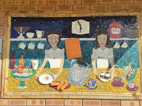 A colourful café-themed mosaic on the Cummins Mosaic Trail, showing two servers with teacups and cak