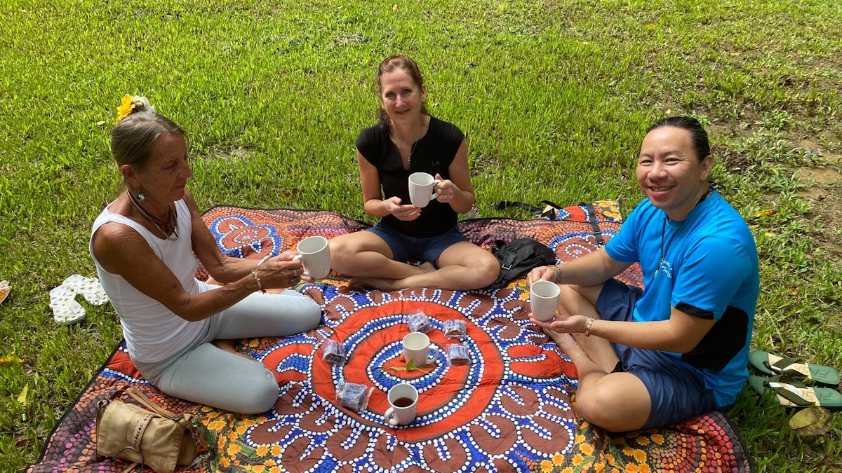 Three people sitting on picnic rug with tea and snacks