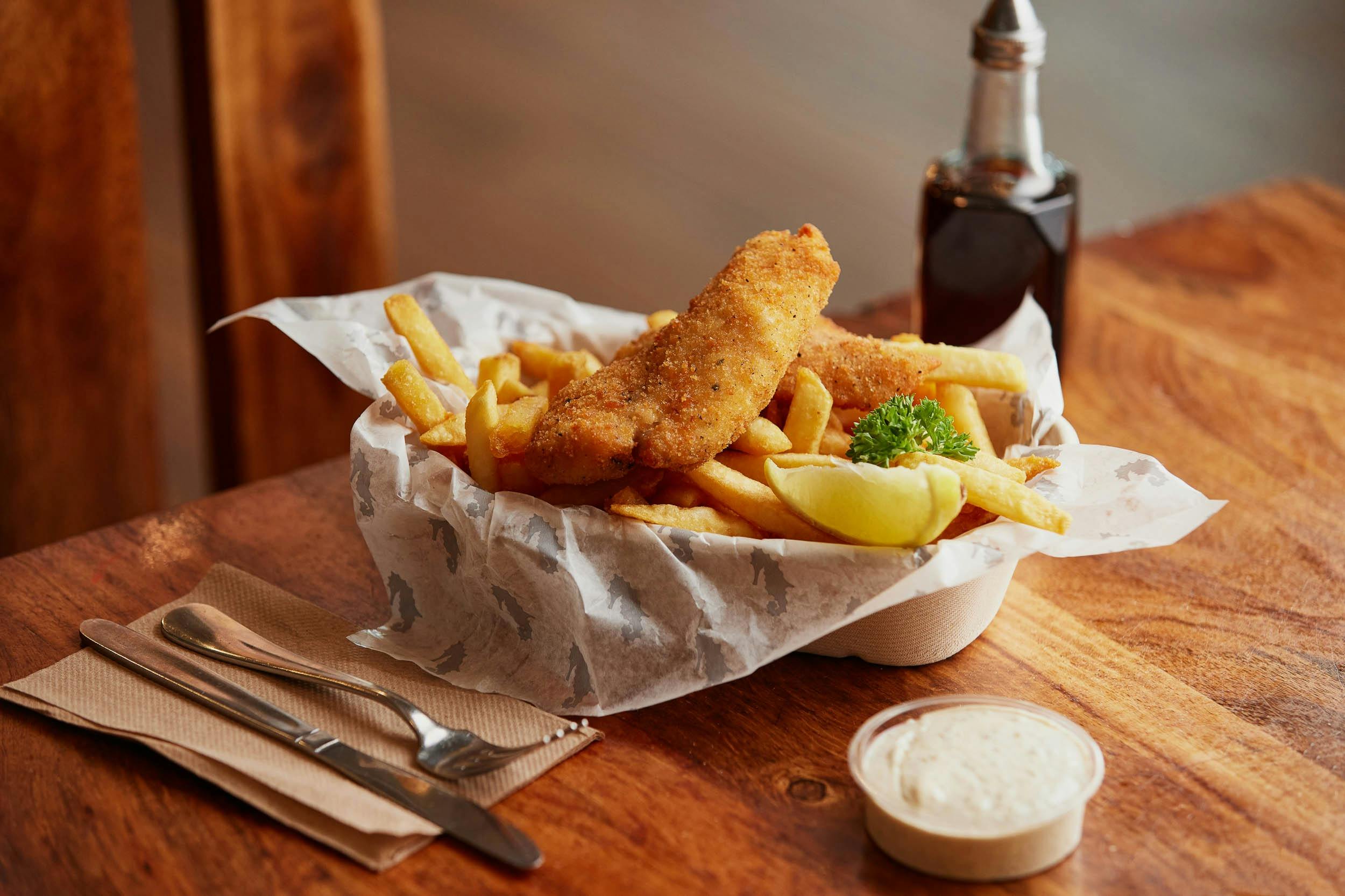 A basket containing fish and chips with tartare sauce and vinegar on the side