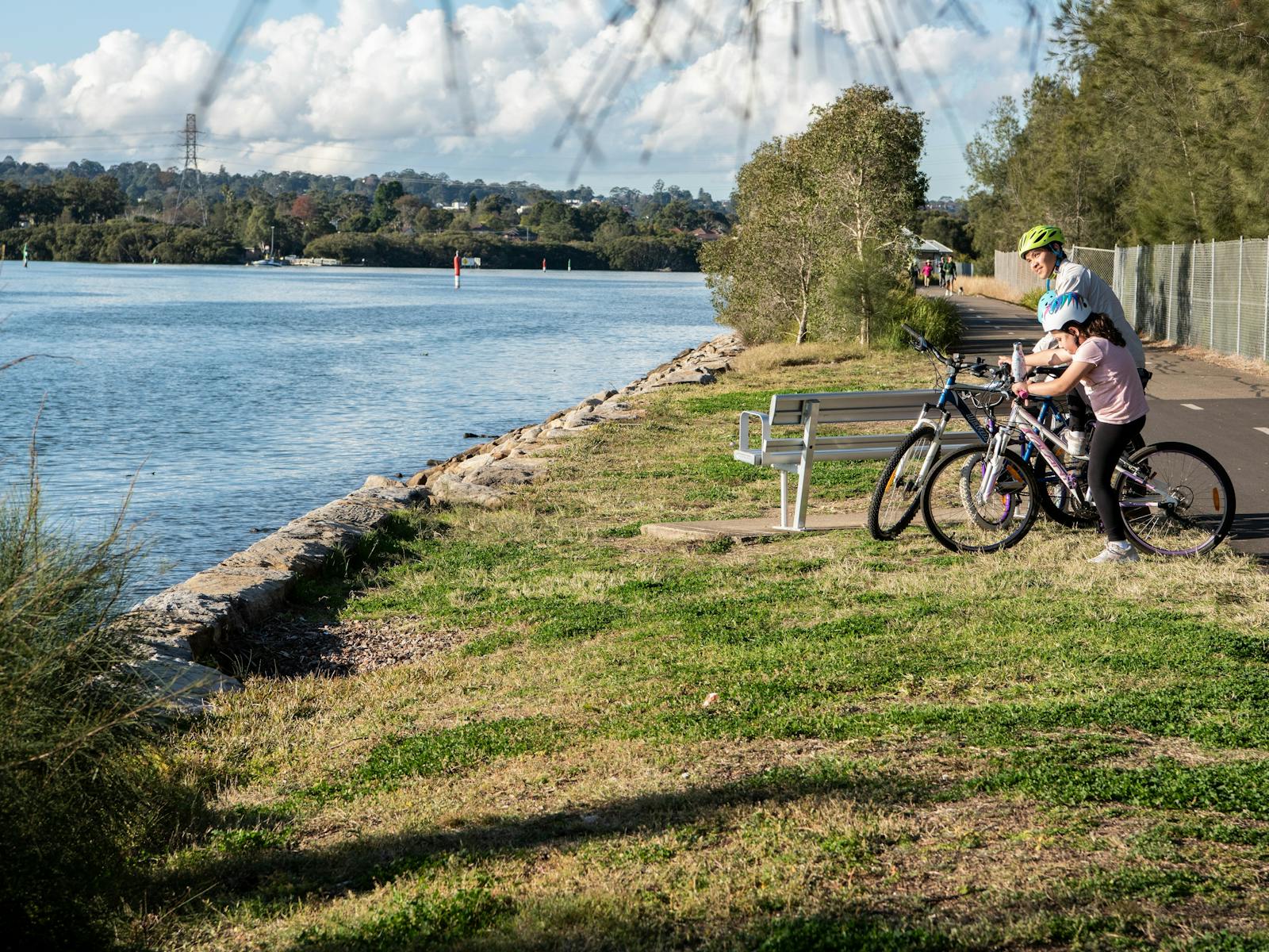 3 bike riders stopped by the river to look at the view, along a grass edge and pathway.