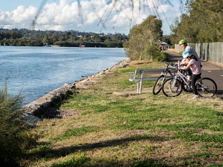 3 bike riders stopped by the river to look at the view, along a grass edge and pathway.