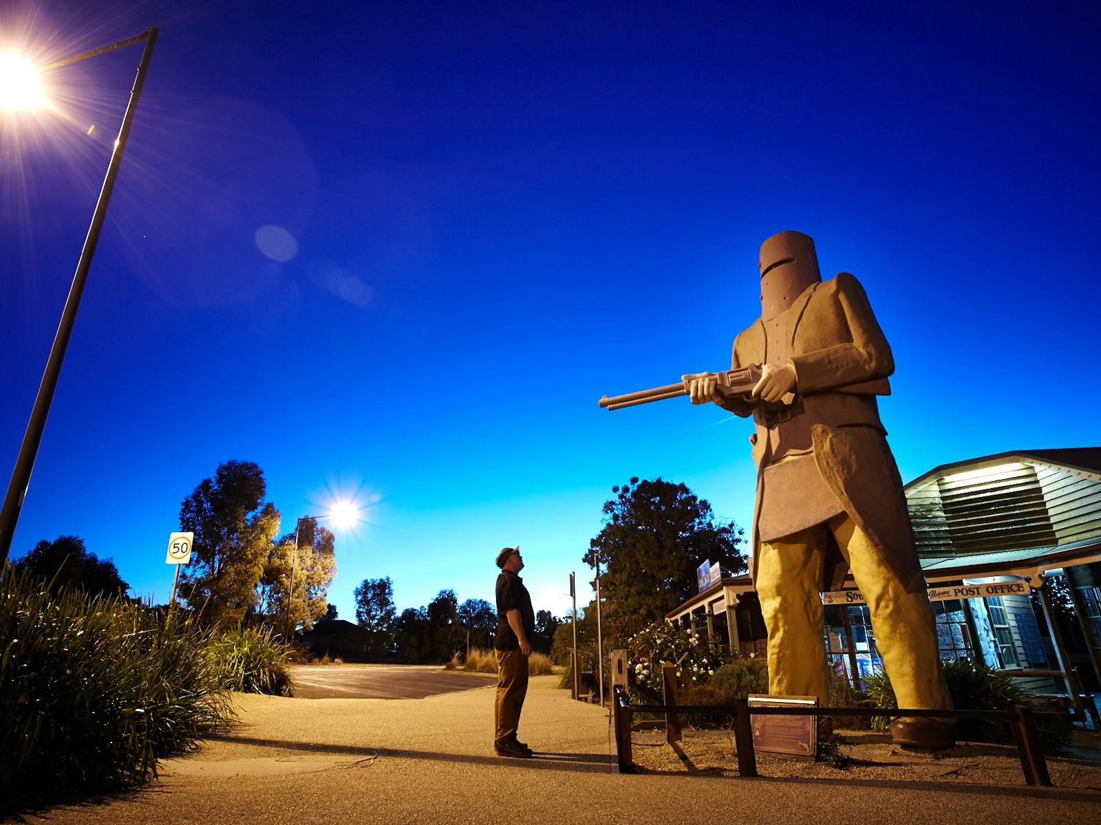 Man standing in front of Ned Kelly Statue at sunset, street lights on