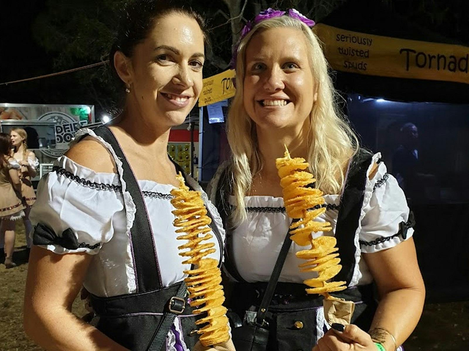 Two girls in traditional Bavarian costume eating spiral potatoes on a stick.