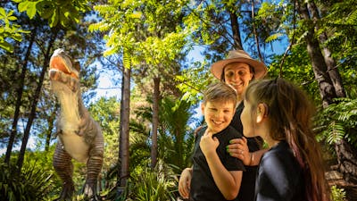 A mother and two children smile in awe at the size of the animatronic T-Rex in lush fern gardens.