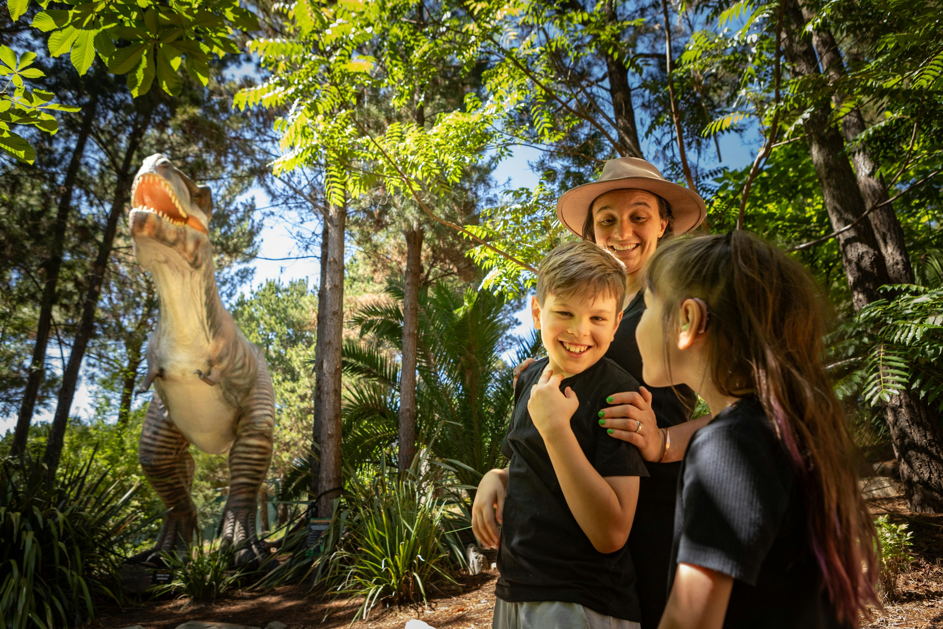 A mother and two children smile in awe at the size of the animatronic T-Rex  in lush fern gardens.