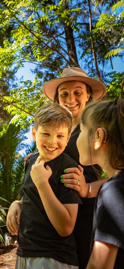 A mother and two children smile in awe at the size of the animatronic T-Rex in lush fern gardens.