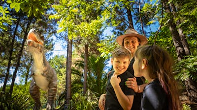A mother and two children smile in awe at the size of the animatronic T-Rex in lush fern gardens.