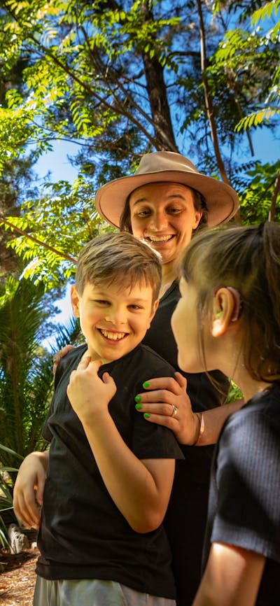 A mother and two children smile in awe at the size of the animatronic T-Rex in lush fern gardens.