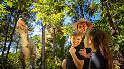A mother and two children smile in awe at the size of the animatronic T-Rex in lush fern gardens.