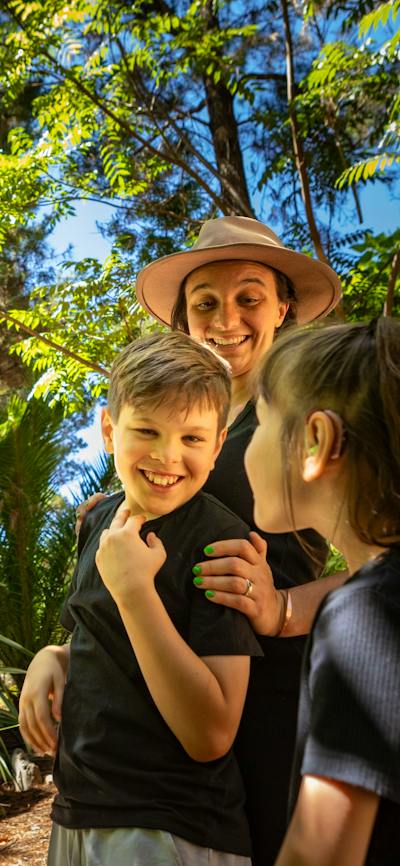 A mother and two children smile in awe at the size of the animatronic T-Rex in lush fern gardens.