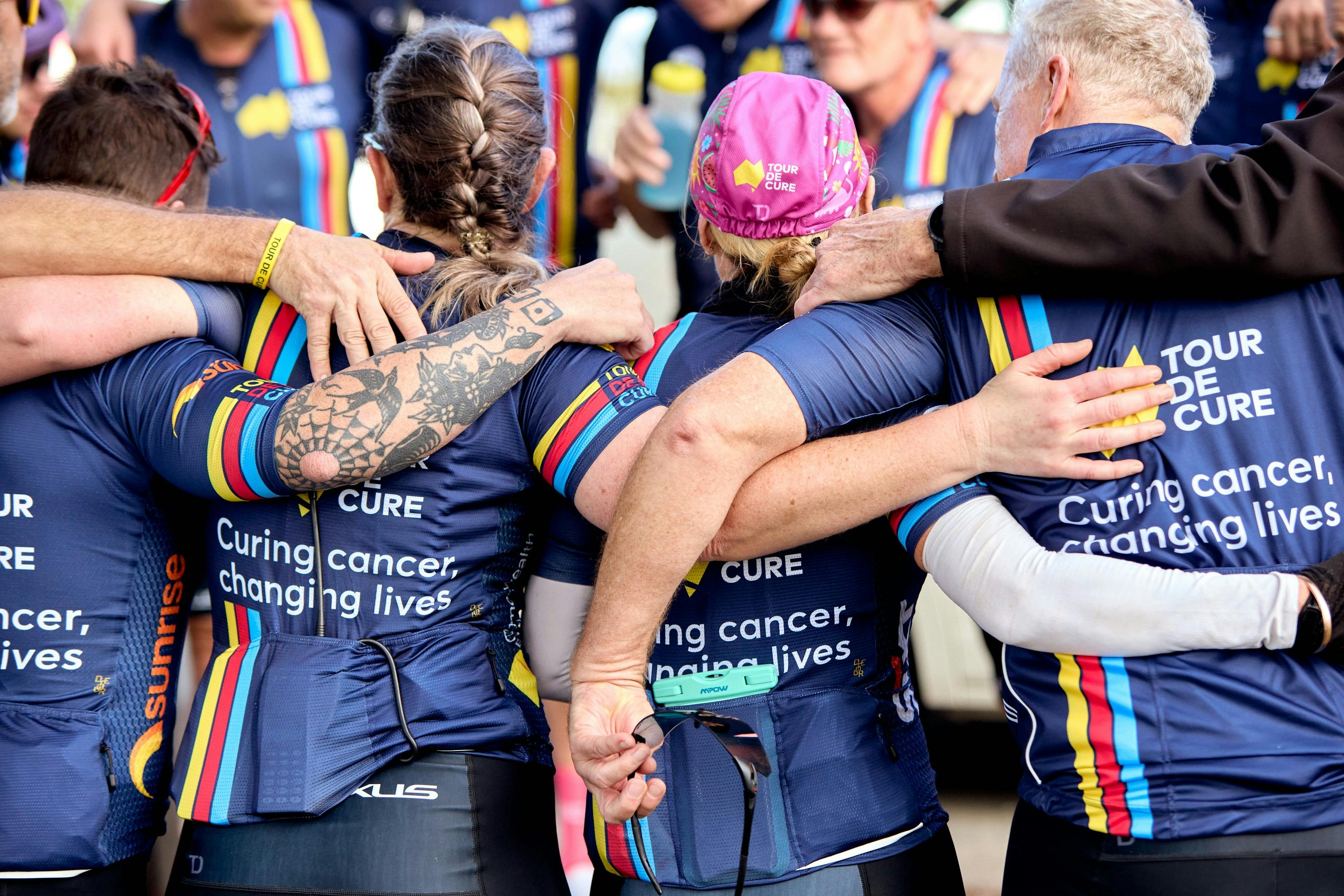 Signature Tour cyclists in kit, in a huddle with arms around each other,  taken from behind them