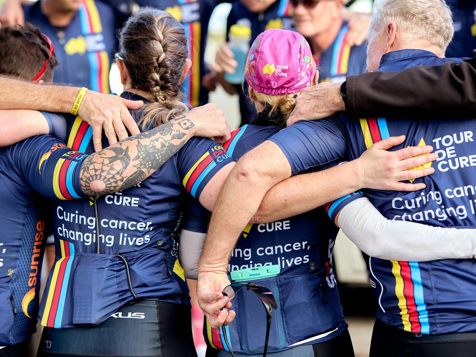 Signature Tour cyclists in kit, in a huddle with arms around each other,  taken from behind them