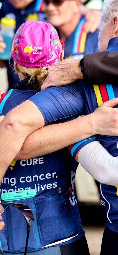 Signature Tour cyclists in kit, in a huddle with arms around each other, taken from behind them