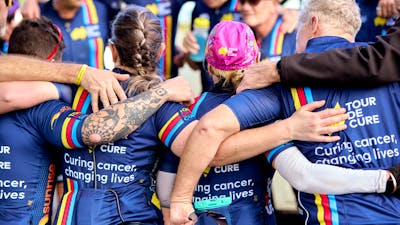 Signature Tour cyclists in kit, in a huddle with arms around each other, taken from behind them
