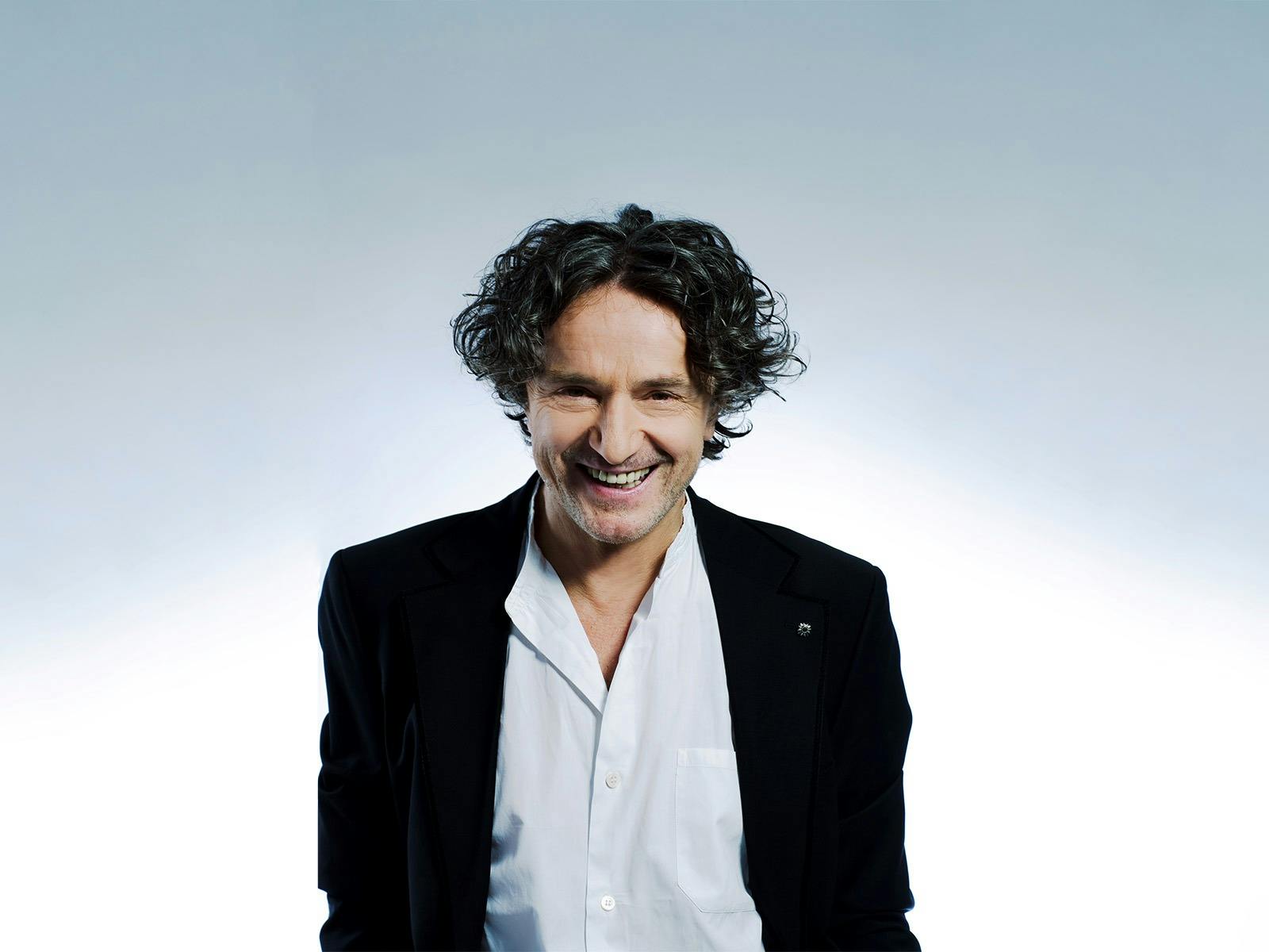 Goran standing in a white button up shirt with a black blazer, in front of a light blue background.
