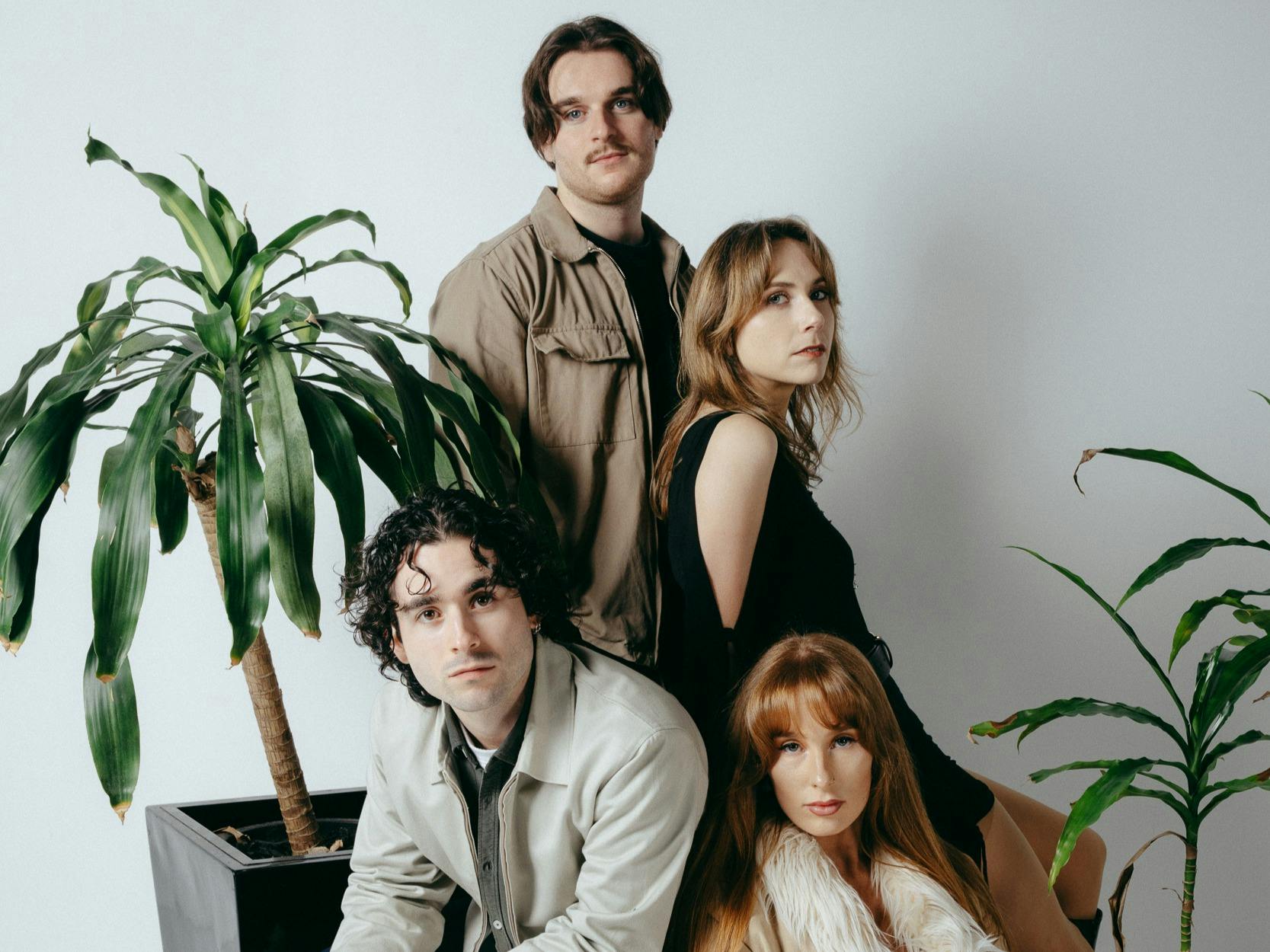 Four band members posed in a cluster among leafy green plants against a white wall.