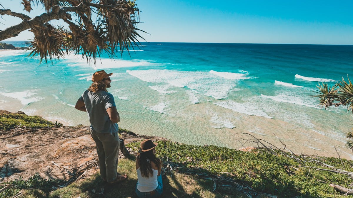 Couple under pandanus tree on cliff, enjoying sunny ocean views on Minjerribah