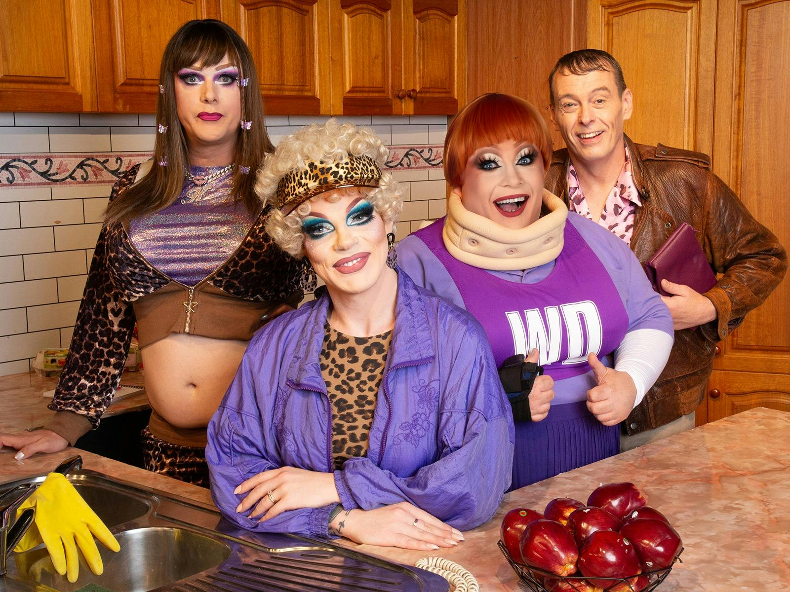 Four performers in colourful Y2K-inspired costumes and theatrical makeup pose together in a kitchen,