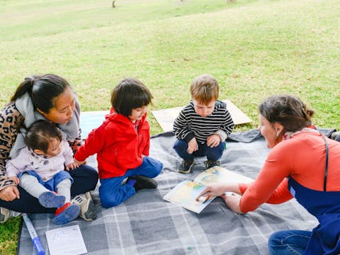 Three toddlers and carer sitting on a picnic rug reading a children's book with a Bundanon educator