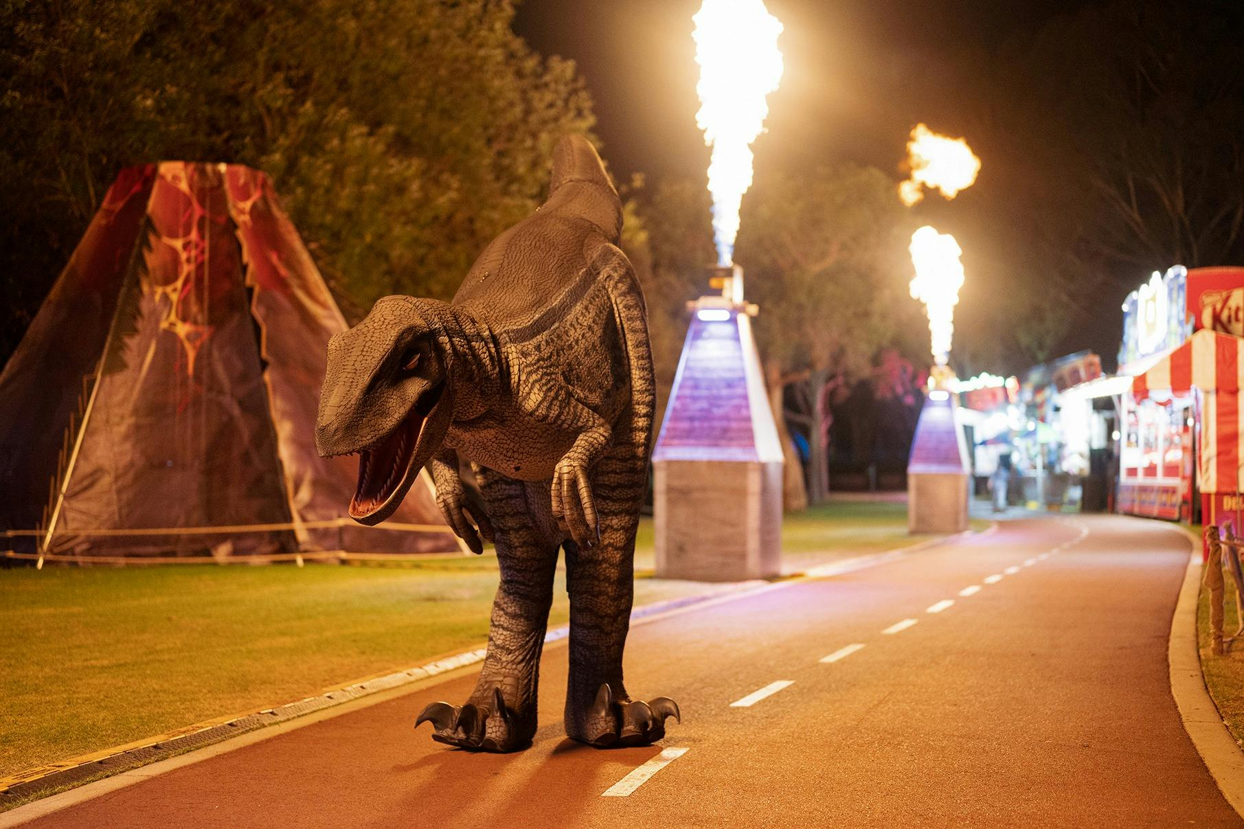 A life-size animatronic dinosaur walking along a footpath, with flame towers in the background.