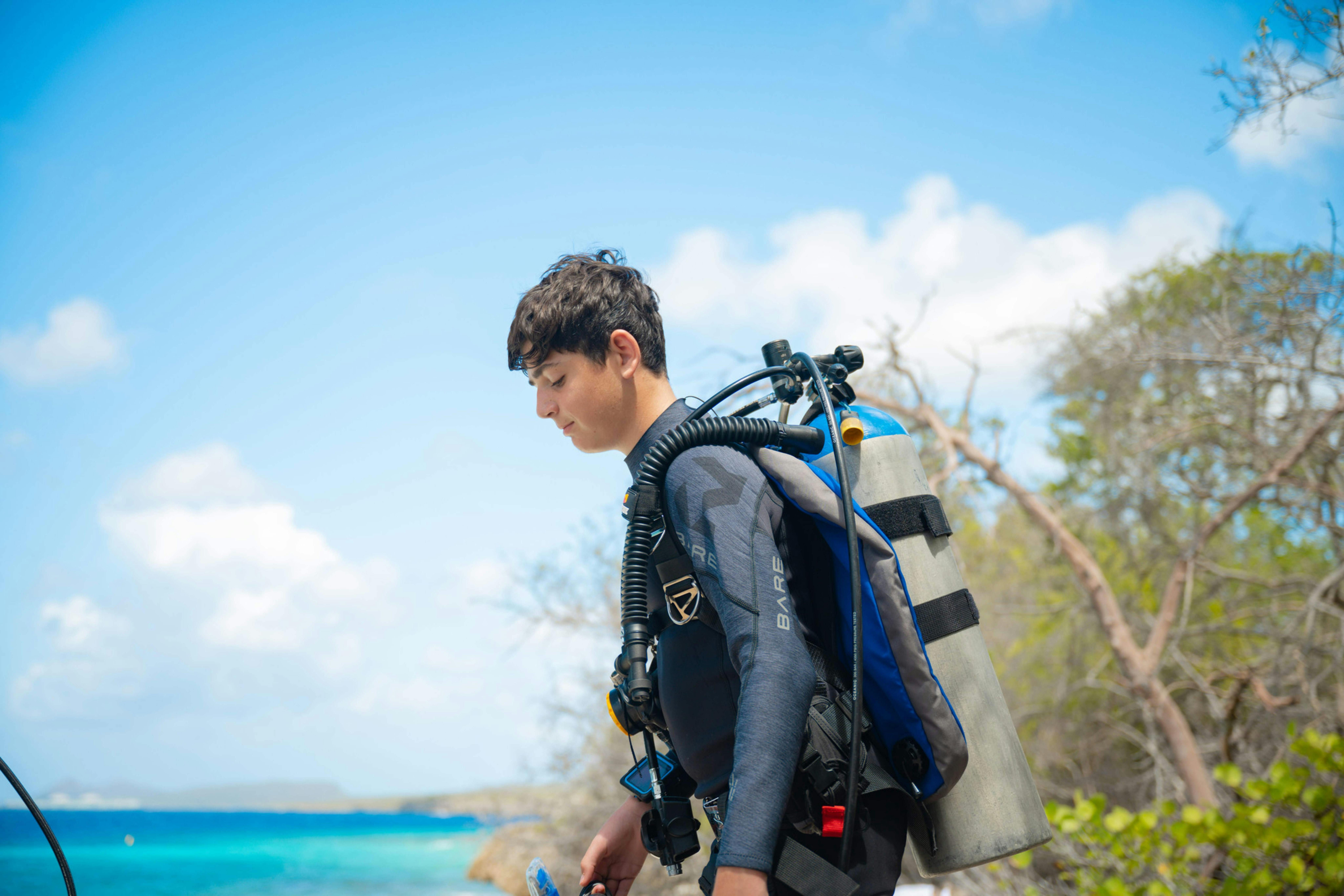 Young scuba diver wearing a wetsuit and full scuba equipment walking towards clear blue water.