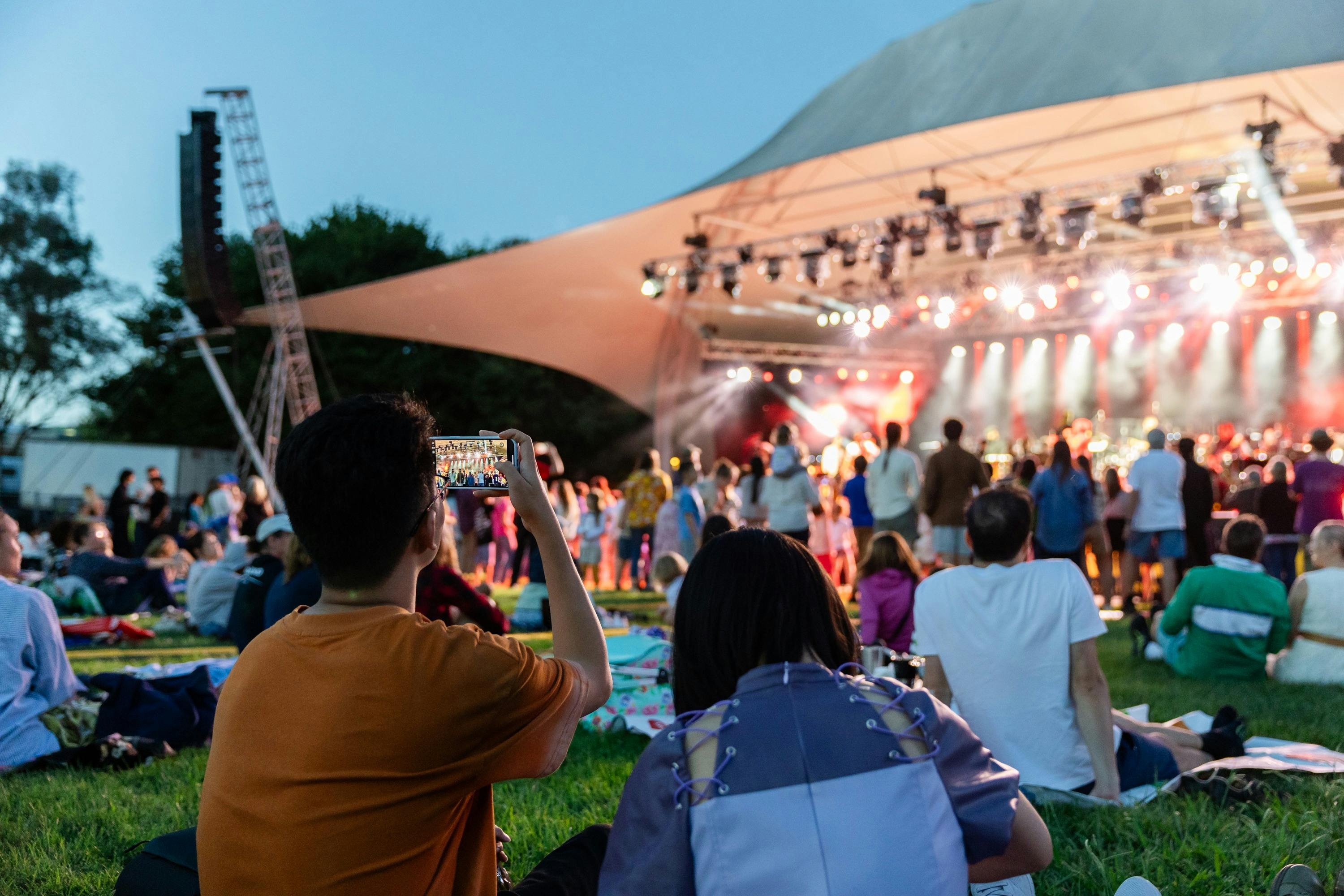 People sitting on grass, looking up at stage as they watch outdoor performance