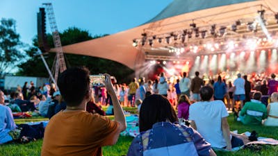 People sitting on grass, looking up at stage as they watch outdoor performance