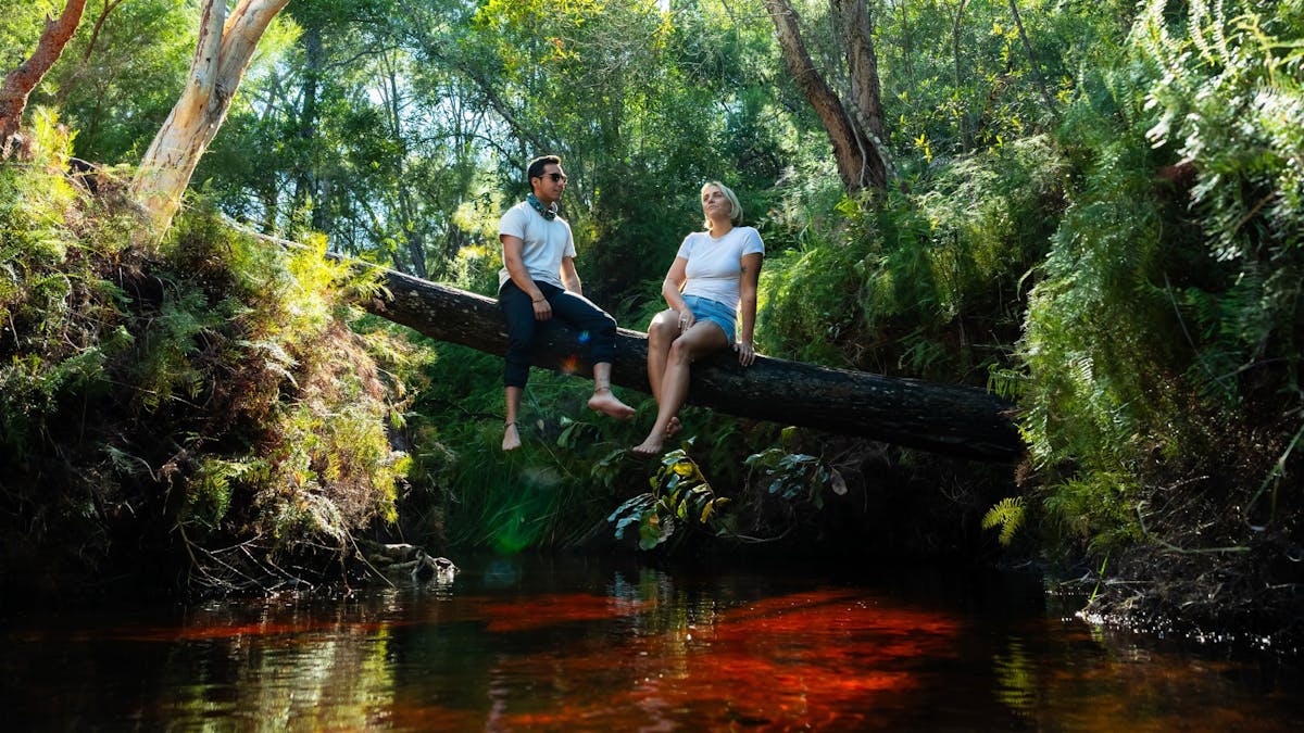 Two people sitting on a long tree branch with the red coloured water underneath with greenery around