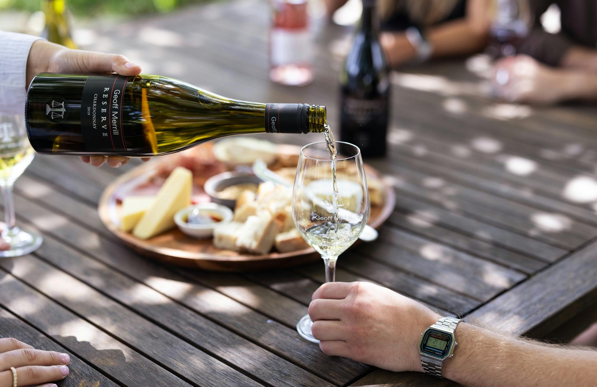 A glass of white wine being poured in fornt of a big cheeseboard