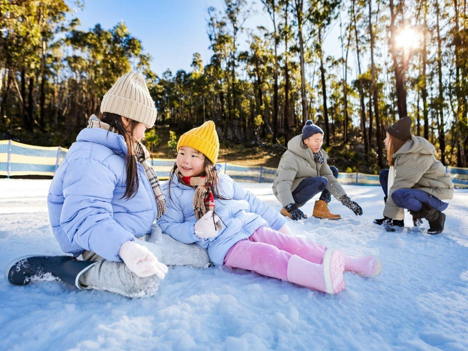 Two girls sit in the snow, smiling at eachother. Their parents crouch behind them, gathering snow