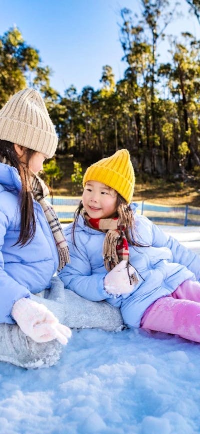 Two girls sit in the snow, smiling at eachother. Their parents crouch behind them, gathering snow