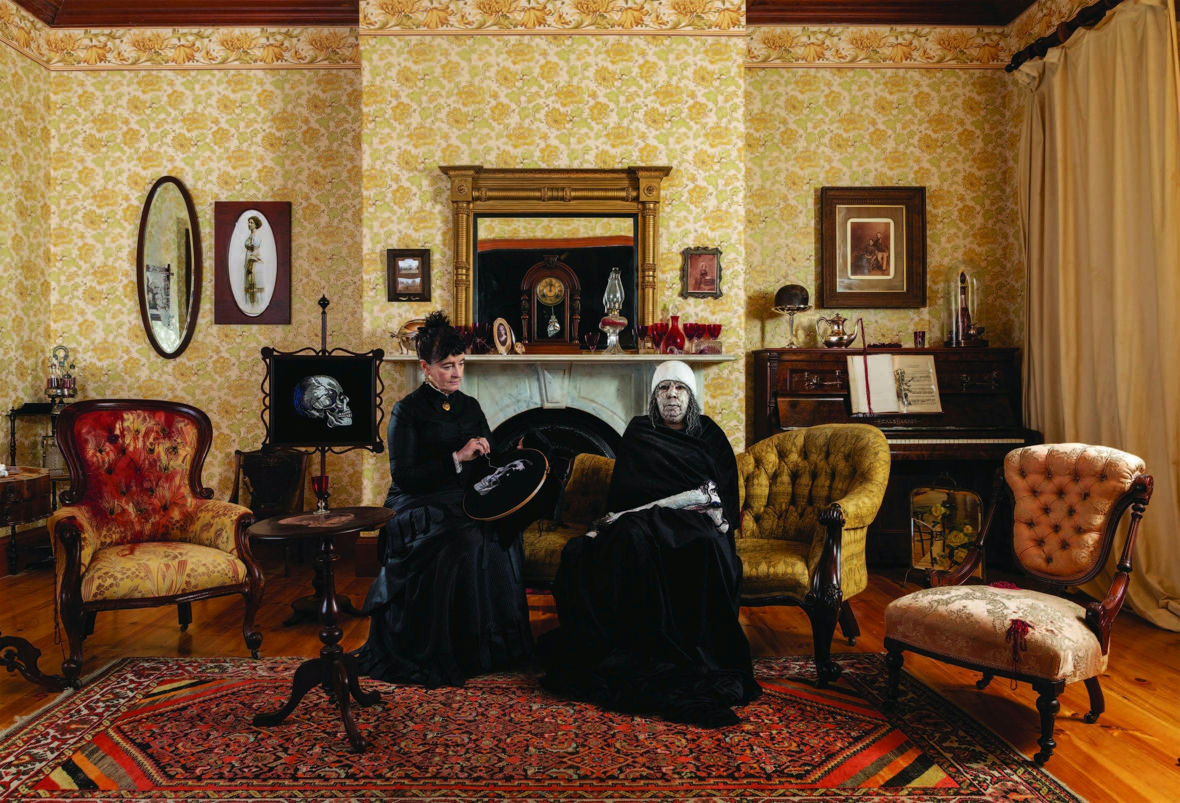 A staged Victorian-style interior, two women dressed in black sit together