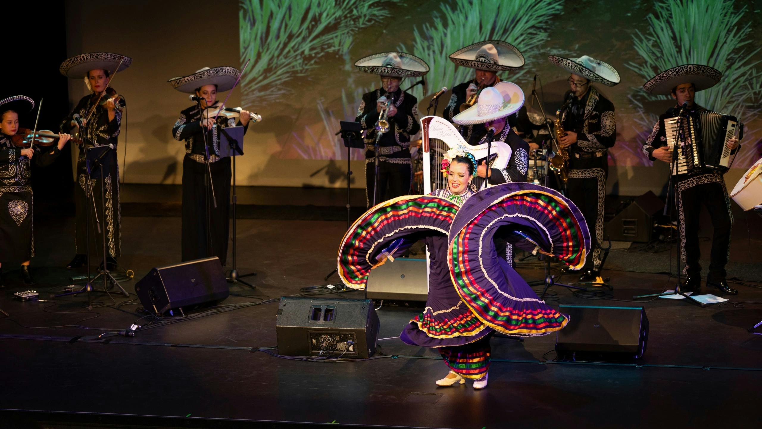 A woman dances on stage in a colourful dress, with a mariachi band performing behind her