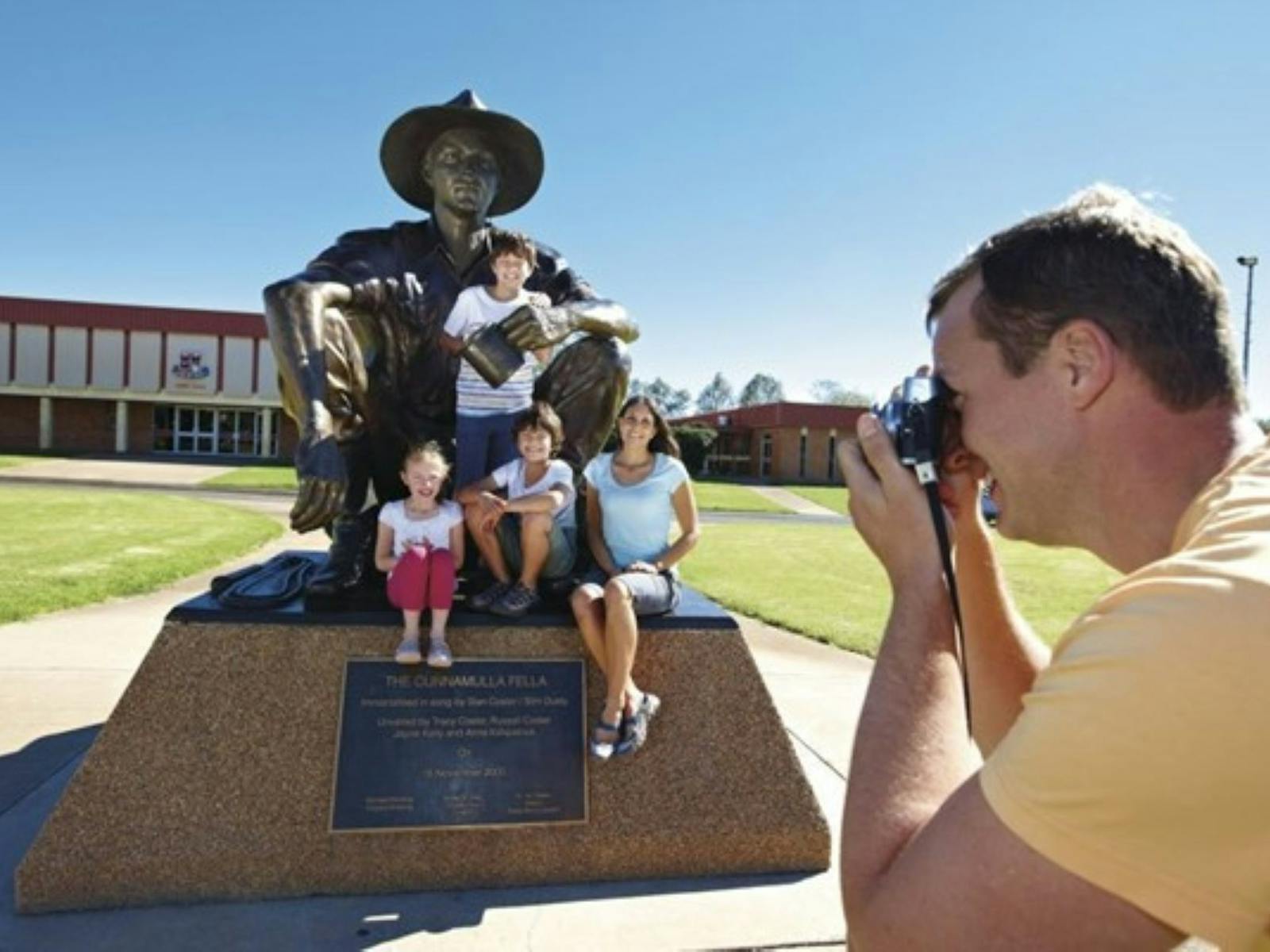 Man taking a photo of family sitting on the Cunnamulla Fella Statue Man taking a photo of family sitting on the Cunnamulla Fella Statue