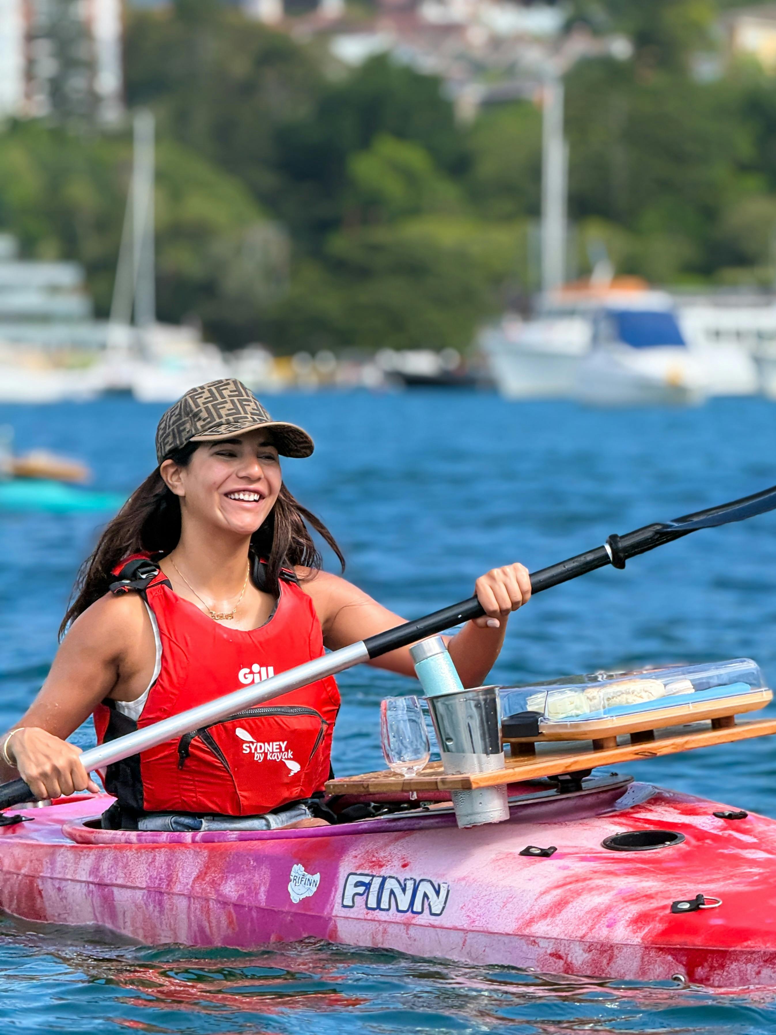 Lady paddling with scones and tea on the front of her kayak smiling as she enjoys Sydney harbor