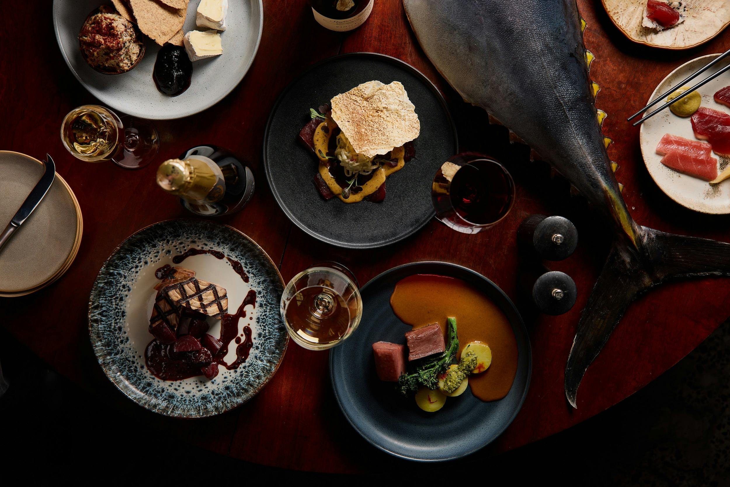 A whole tuna tail sit on a table, surrounded by restaurant dishes created from tuna.