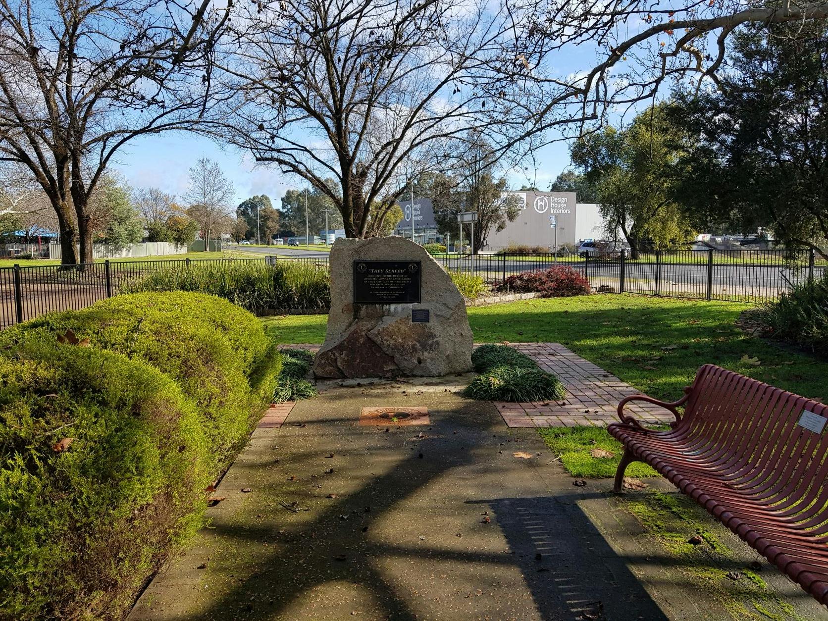 Shrubs, seat, rock with memorial plaque, paving bricks, fencing, trees, road