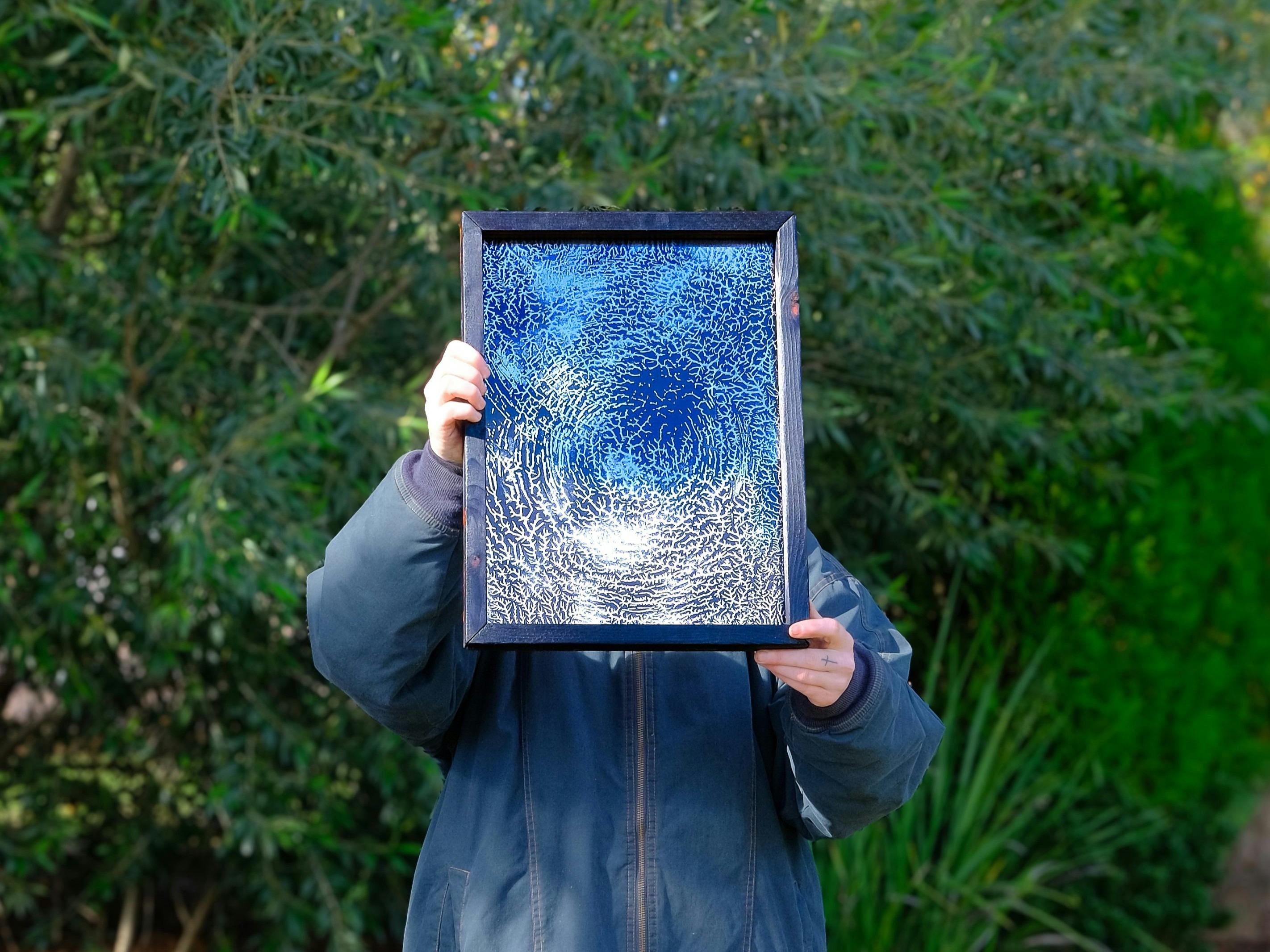 The artist holds a framed artwork of an etched mirror in front of their face, blocking it.