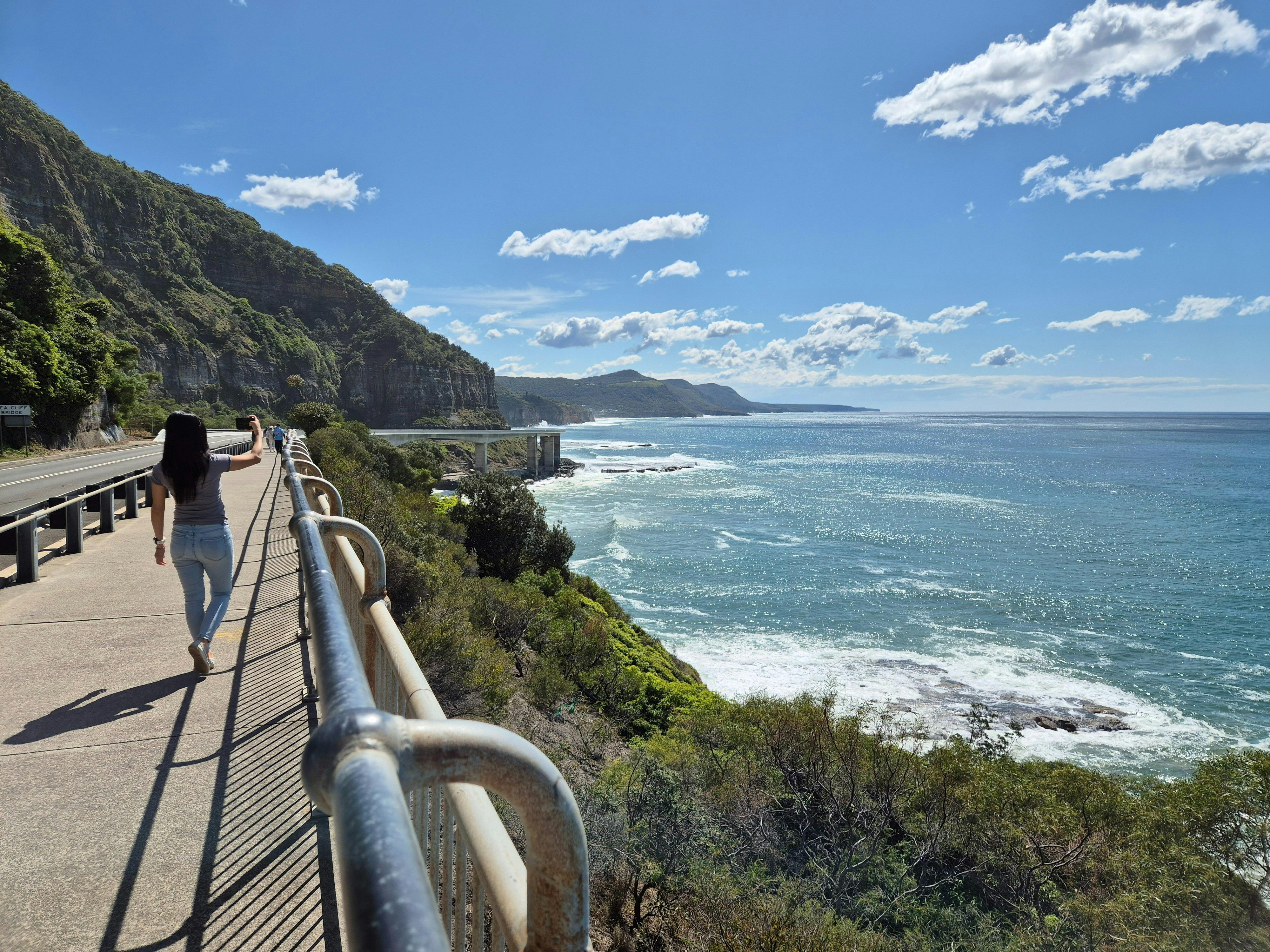 Person walks along Sea Cliff Bridge on a sunny, blue sky day