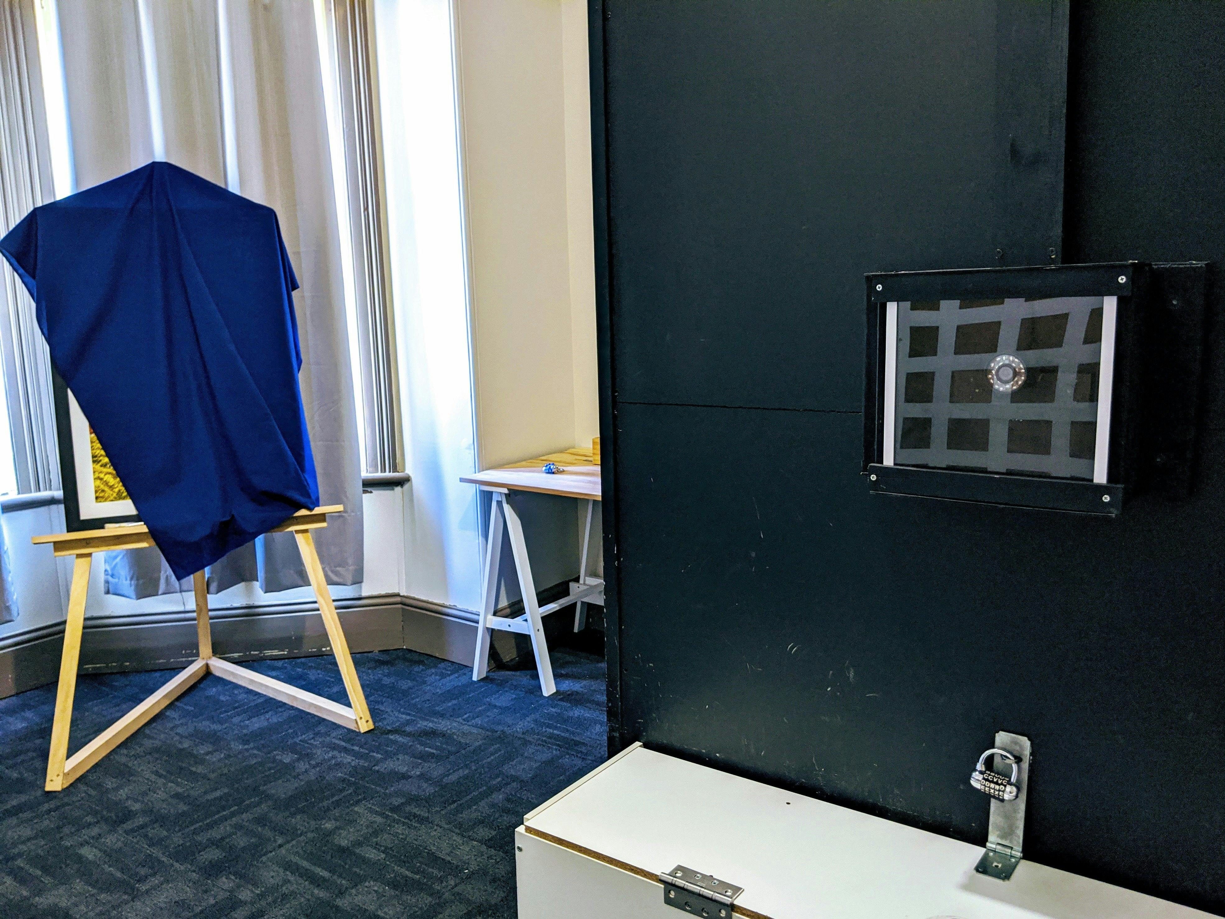 Black wall with camera mounted inside a box with a fabric covered easel and desk in the background