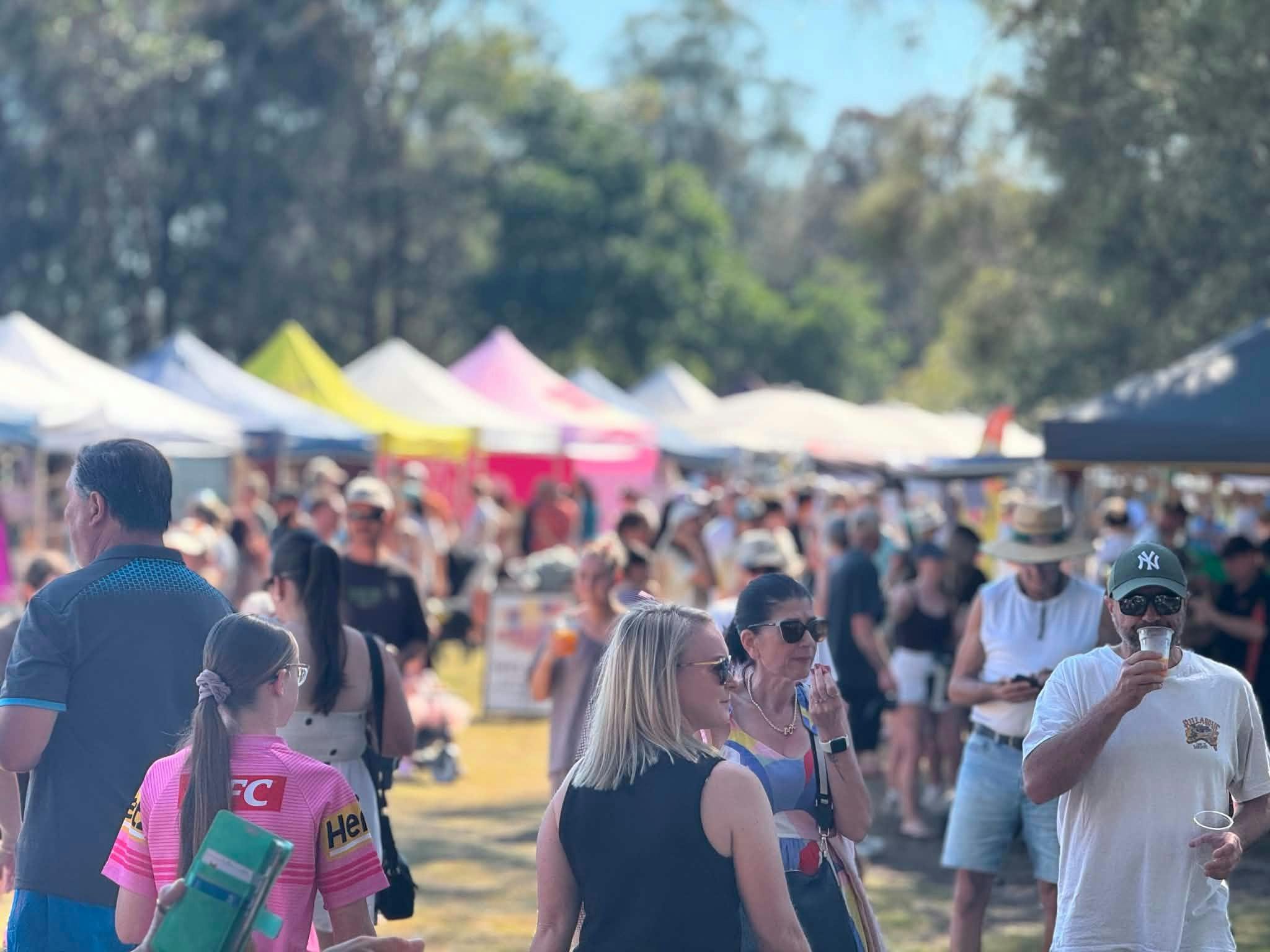 Crowds exploring colourful market stalls beneath gazebos and trees on a sunny day.
