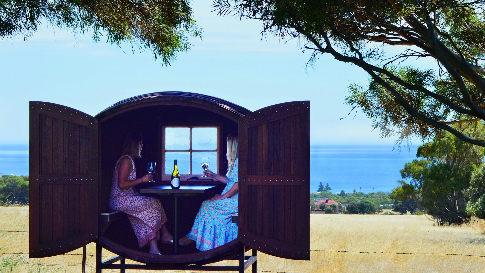 Two ladies drinking wine inside a giant wine barrel tasting pod, overlooking sea views