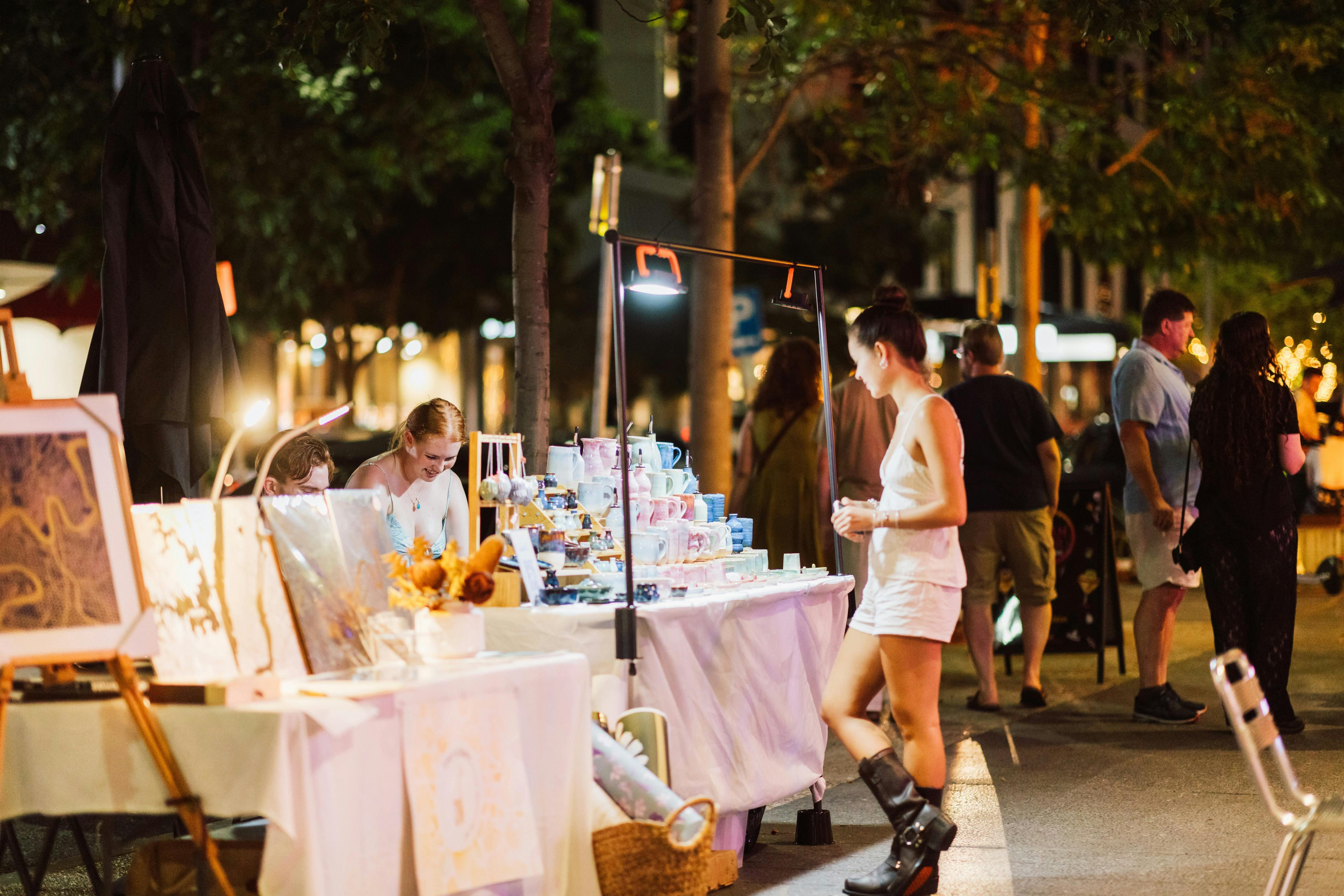 Woman browses handmade ceramics and artwork at a warmly lit outdoor night market.
