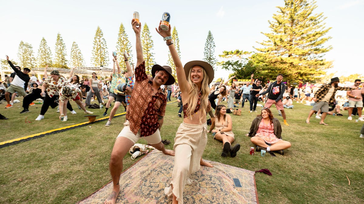A couple on a picnic rug doing a yoga pose holding beers to the sky