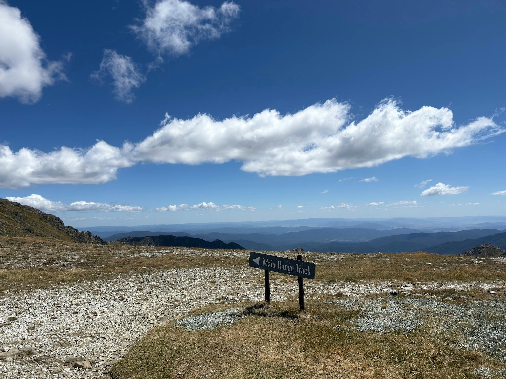 A sign on a hiking trail with blue skies and clouds in the background.