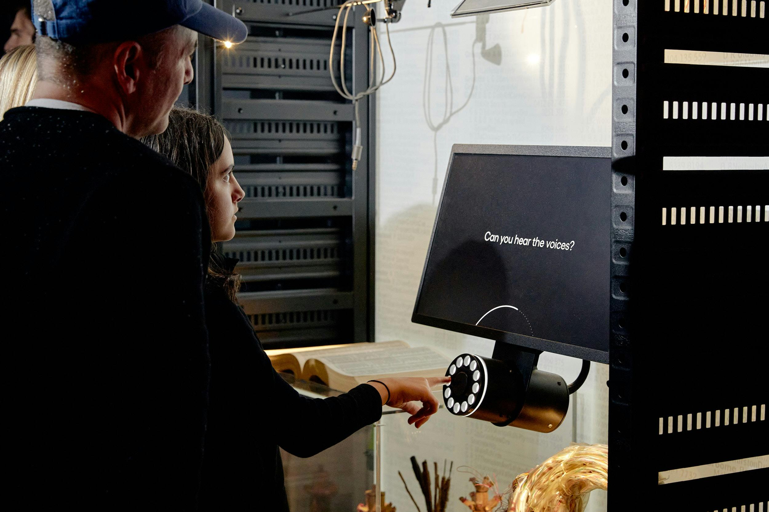 A woman interacts with a rotary dial. The screen reads "Can you hear the voices?"