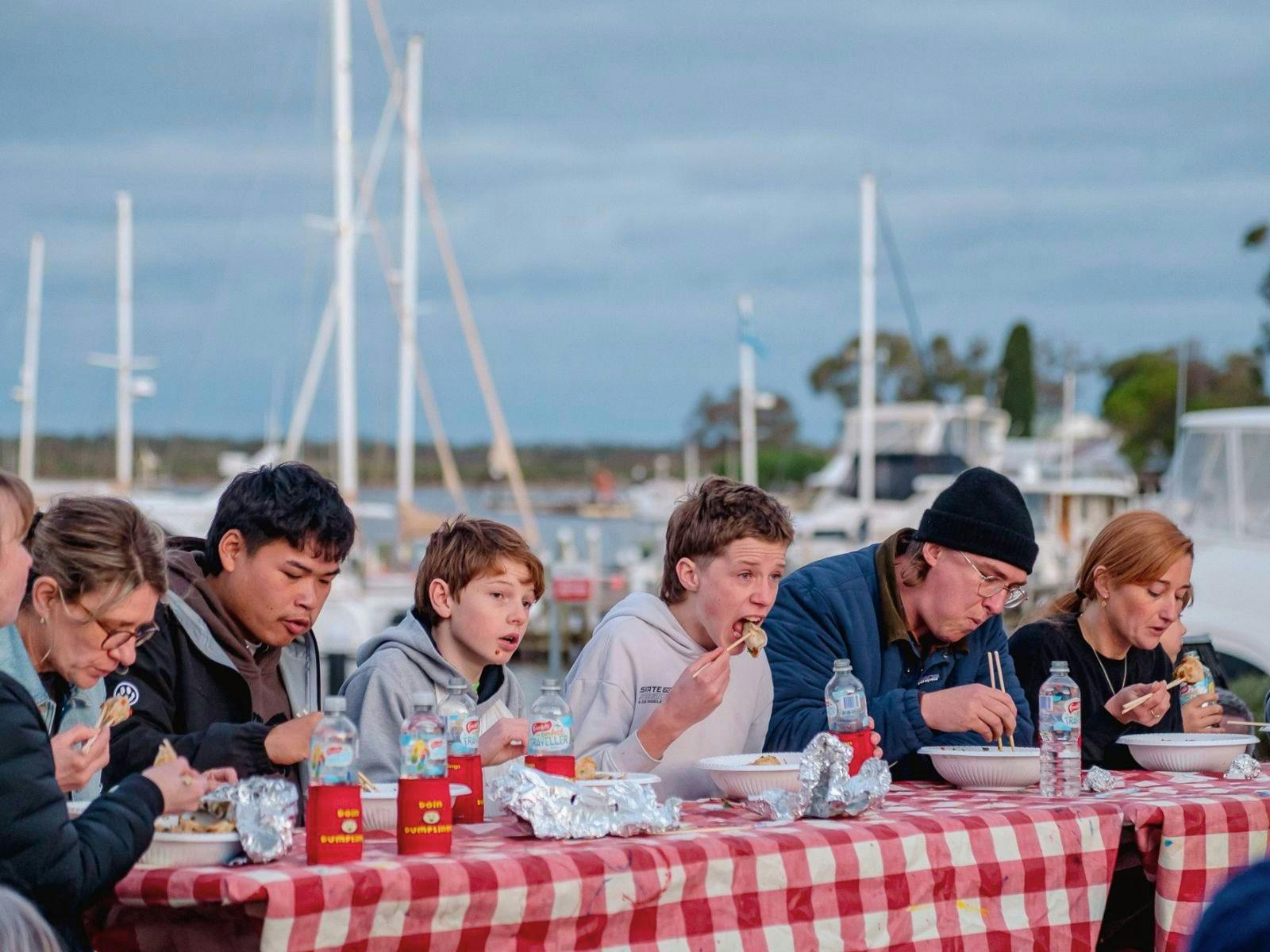 Row of people sitting a table eating dumplings with lake and yachts behind